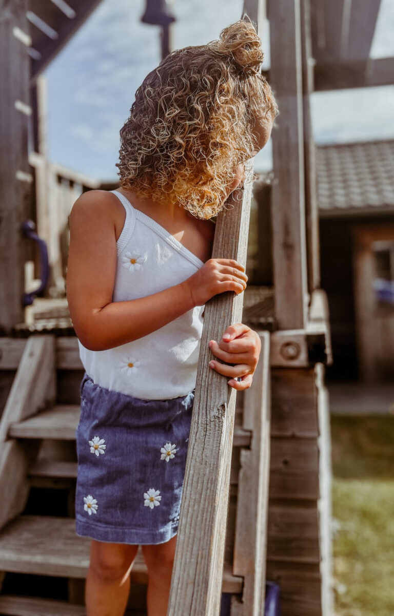 Meisje met blonde krullen in een wit topje met madeliefjes, leunend tegen een houten speeltoestel