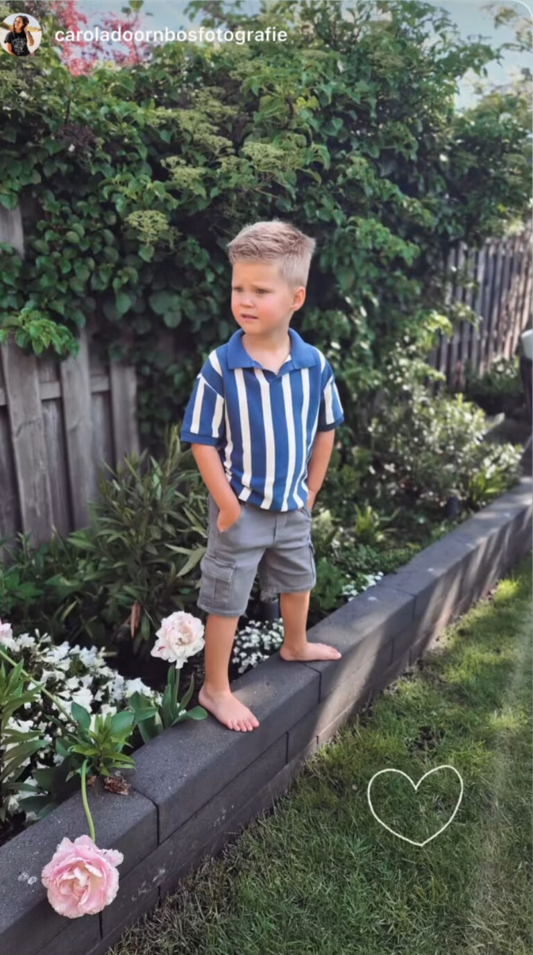 Jongen in een blauw-wit gestreept poloshirt en grijze korte broek, blootsvoets op een tuinrand tussen de bloemen