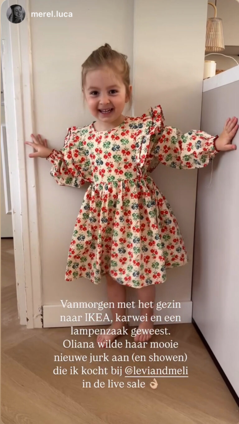 Meisje in een crèmekleurige jurk met ruches en rood-groene bloemenprint, stralend in de gang thuis