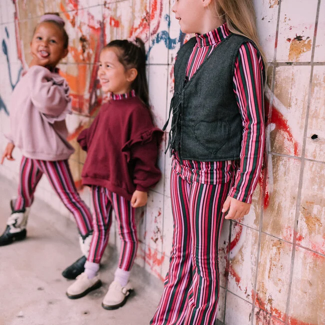 Kinderen dragen Jacky Pants Funky Stripe broek in roze en zwarte strepen gecombineerd met sweaters en gilet.